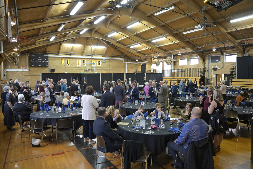 People gathered in a gymnasium for an event, socializing around decorated tables with blue napkins and glassware in a community setting. Attendees include adults and children engaging in conversations. The venue features a wooden ceiling structure and sports equipment.