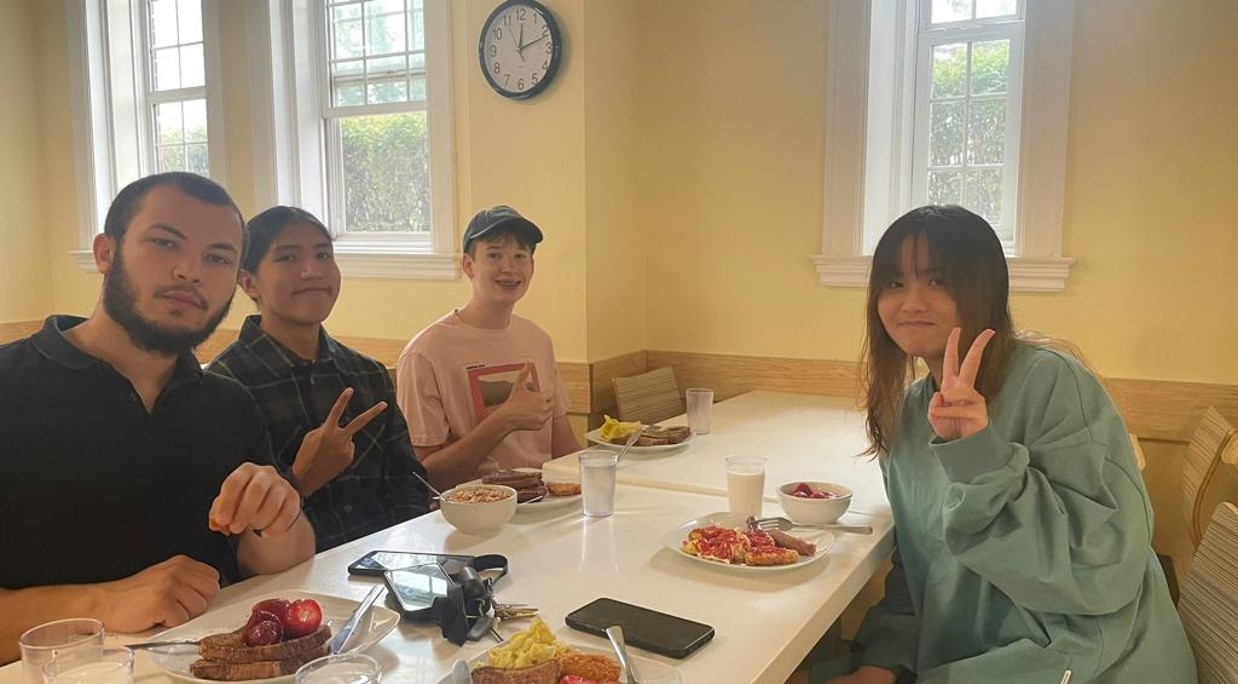 Students Enjoying Breakfast in a Cozy Kitchen Setting Four friends smiling and making peace signs while enjoying a hearty breakfast in a cozy kitchen with natural light. Plates filled with toast, fruit, eggs, cereal, and milk are on the table, creating a warm and inviting atmosphere for friends to gather and start their day.