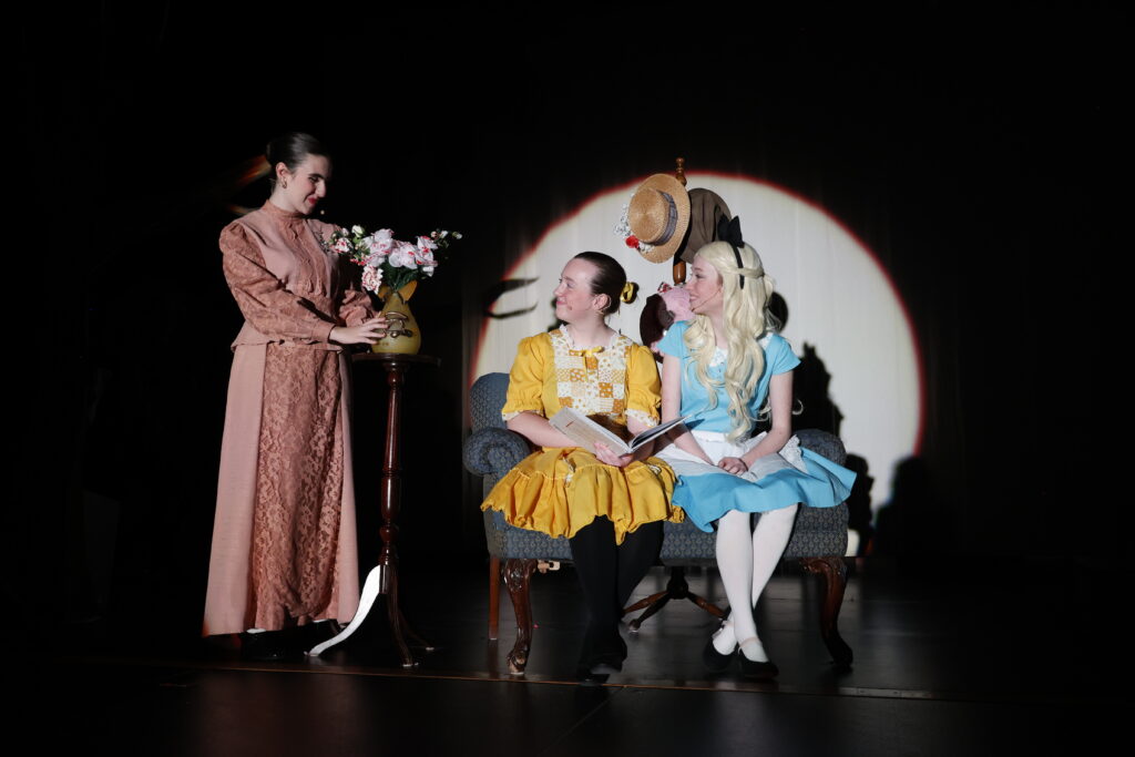 Three actors perform on stage in a theater production. One actor in a long pink dress stands beside a vase of flowers, while two other actors, dressed in yellow and blue period costumes, sit on a couch. The scene is illuminated by a spotlight in the dark theater, creating a dramatic and engaging atmosphere. The performance features detailed costumes and set design, enhancing the historical and nostalgic ambiance of the play.