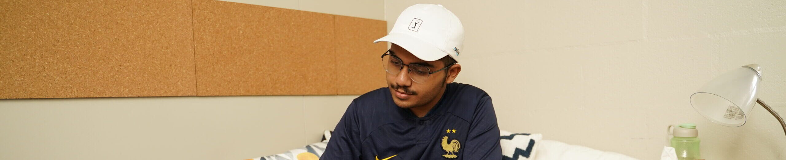 A young man in a blue sports jersey with a white cap and glasses studying in a cozy room with a corkboard wall and a desk lamp.