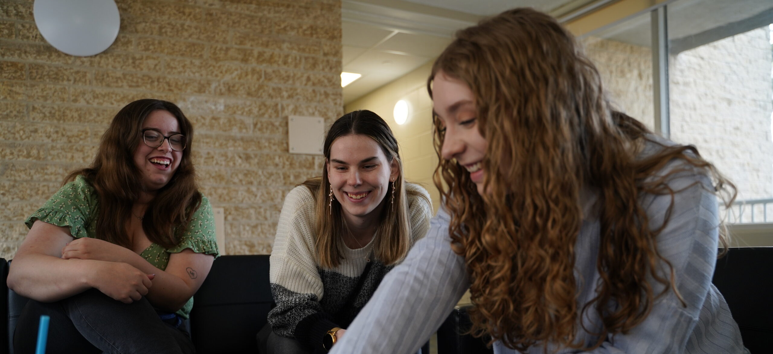 Three young women laughing and smiling while sitting together indoors, creating a joyful and friendly atmosphere.